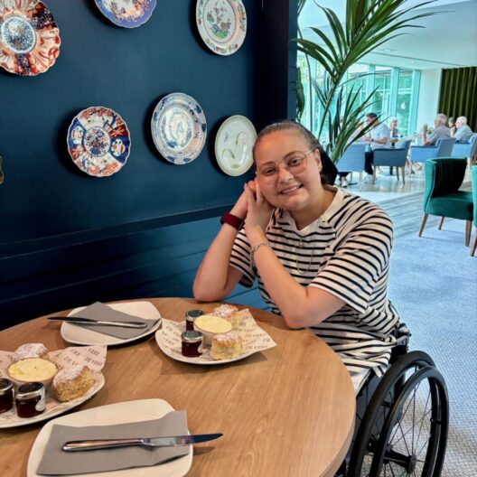 Daisy is sitting in a wheelchair by the table, smiling while having a scone