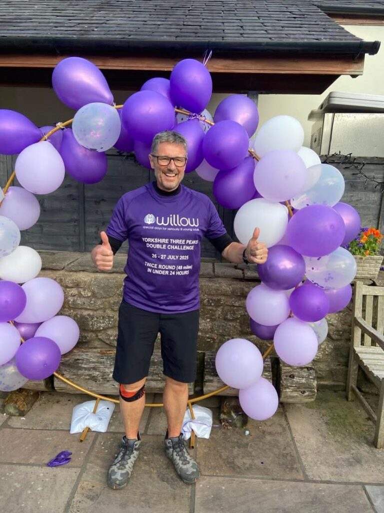 Rob smiles with his thumbs up in front of purple balloon arch to celebrate completing his double three peaks challenge