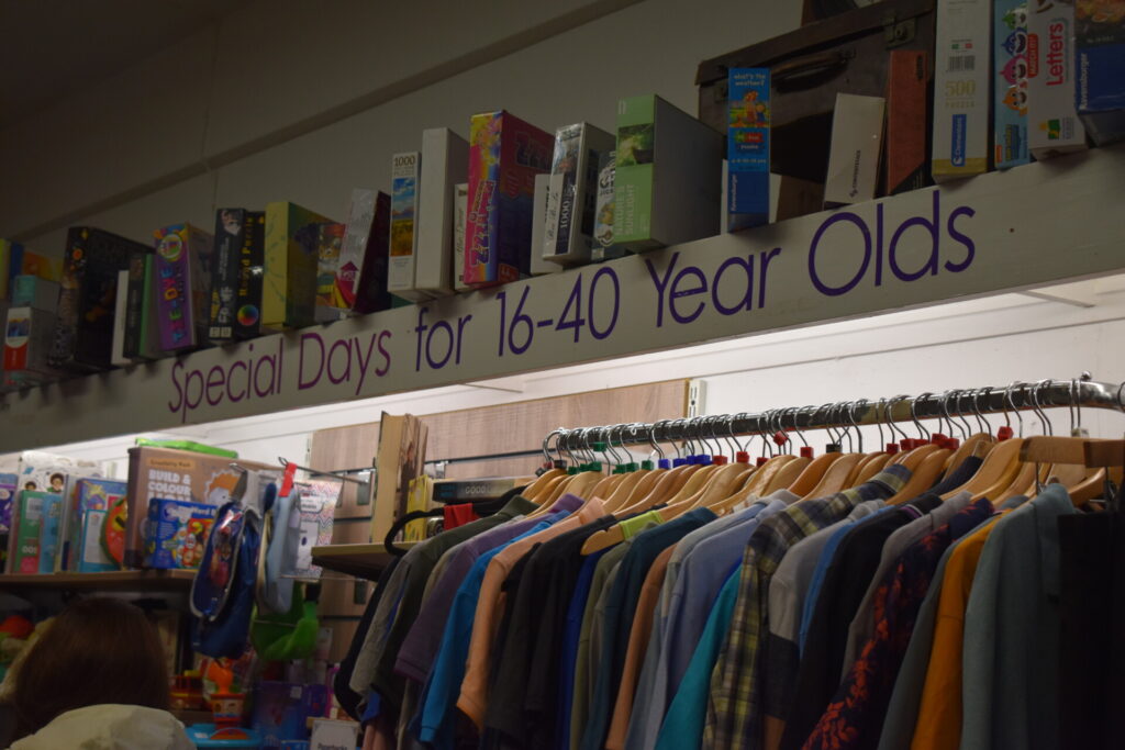 Boardgames sit on shelf above pre-loved clothing rails. Wall stickers read "Special Days for 16-40 Year Olds". 
