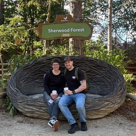 Roxanne sat with her husband sat on a wooden nest, with sign "Center Parcs Sherwood Forest" above them both.