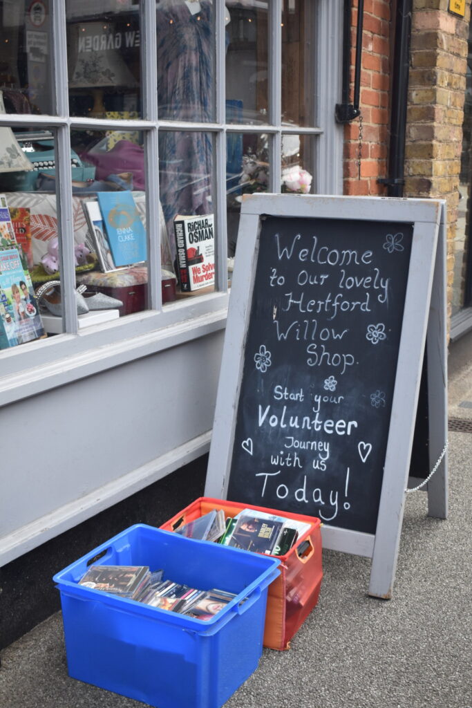 Sign outside reads "Welcome to our lovely Hertford Willow shop. Start your volunteer journey with us Today!"
