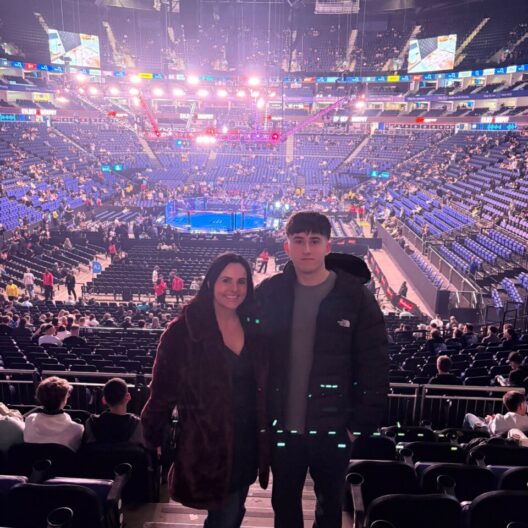 A teenage boy and his mum stand in an arena. There is a UFC ring behind them